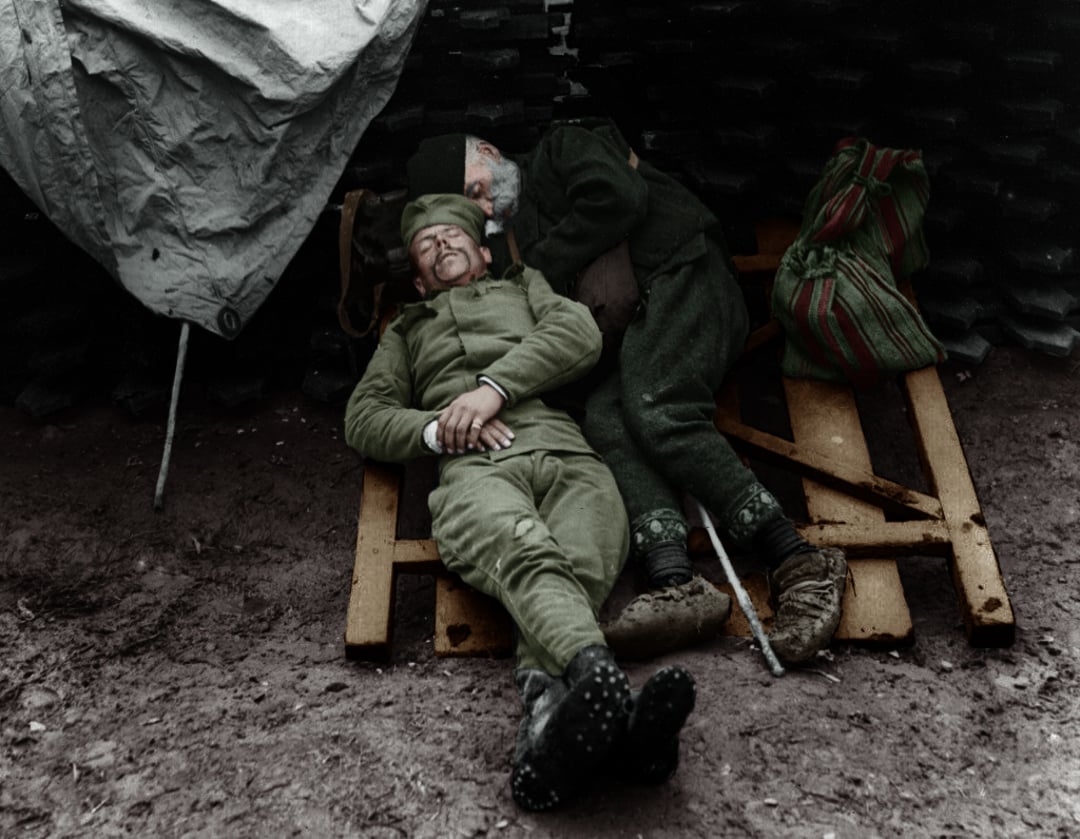 Two soldiers in uniform rest on a makeshift wooden pallet bed on the ground, one lying on his back and the other curled beside him. A striped bag and a cane are placed nearby.