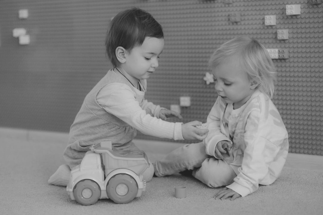 Two young children sit on the floor playing together. One child hands a toy block to the other, who is holding a piece. A toy truck is beside them, and toy blocks are attached to the wall in the background.