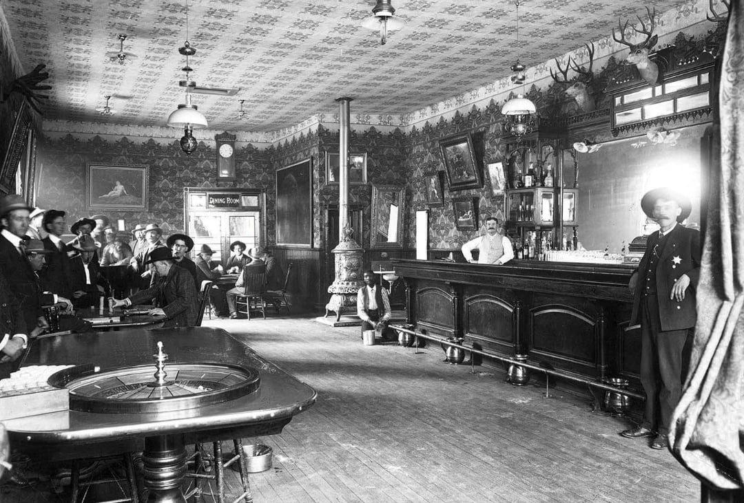 Black-and-white photo of an old Western saloon with men in hats and suits, a bartender behind a long wooden bar, a roulette table in the foreground, and animal mounts and patterned wallpaper decorating the room.