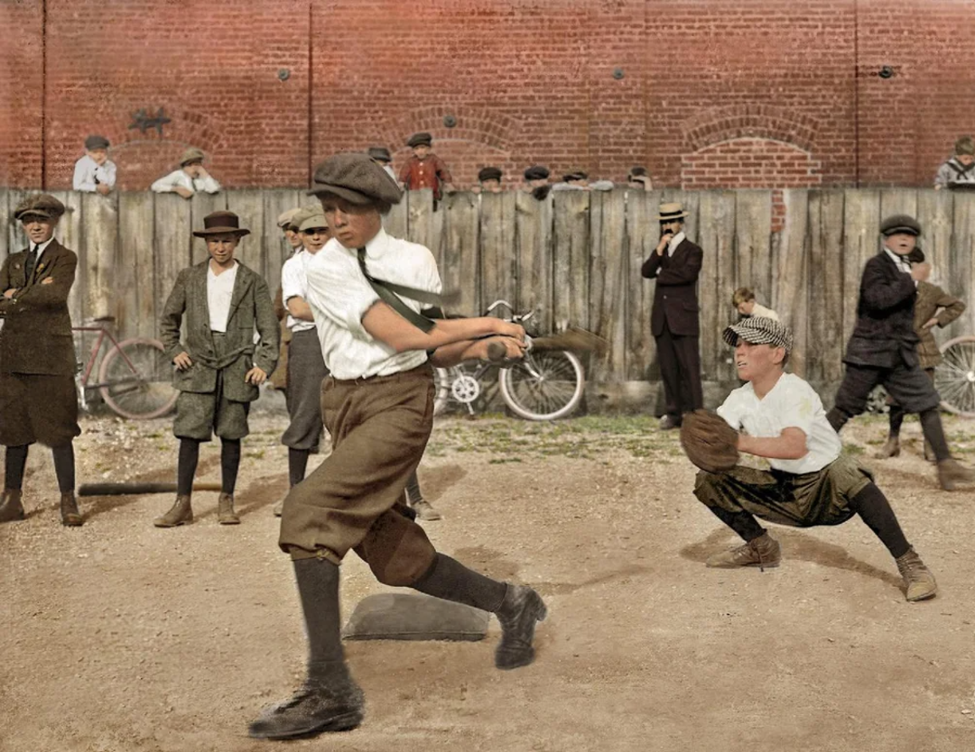 A group of boys in vintage clothing play baseball on a dirt lot, with one batting and another catching. Several bikes and onlookers stand by a wooden fence and brick wall in the background.