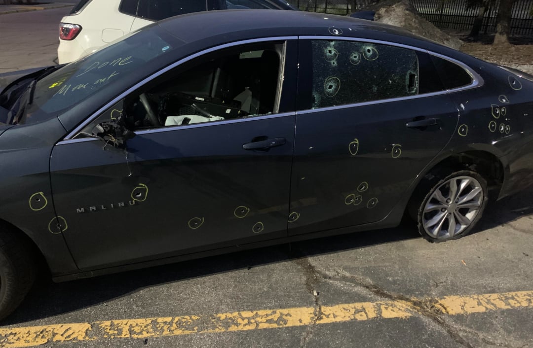 A damaged dark gray Chevrolet Malibu with numerous bullet holes marked in yellow circles along the side, broken windows, and a flat rear tire, parked in a lot near a white SUV.