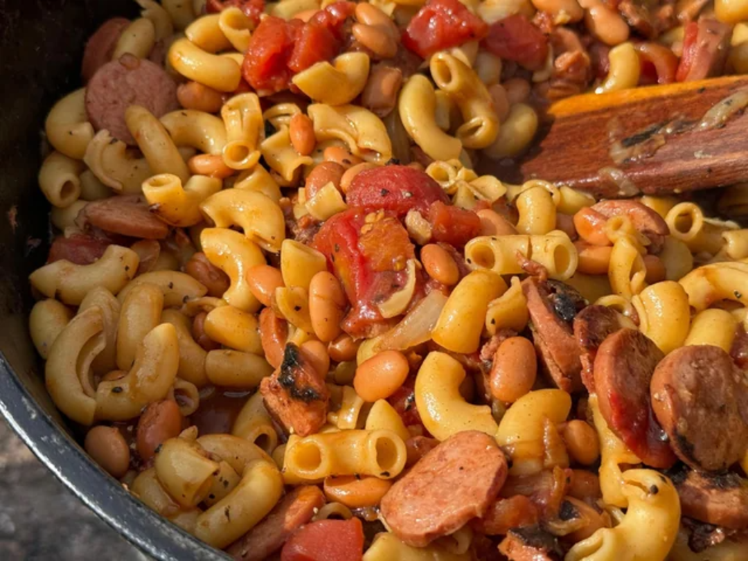A close-up of macaroni pasta mixed with sliced sausage, beans, diced tomatoes, and herbs in a pot, with a wooden spoon partially visible.