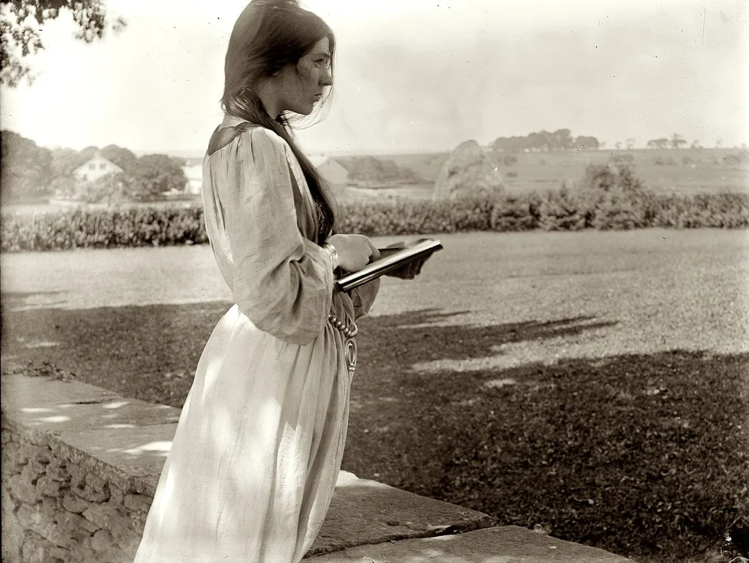 A young woman in a long, light-colored dress stands outdoors by a stone wall, holding an open book and looking thoughtfully into the distance. Trees, fields, and a house are visible in the background.