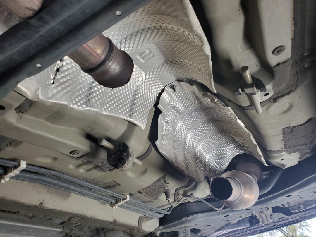 The image shows the underside of a vehicle with a missing catalytic converter, evident by the cut pipes and exposed heat shield where the part has been removed.