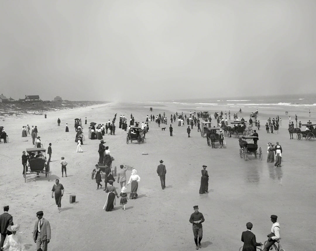 Black-and-white photo of a crowded beach with people in early 20th-century clothing, horse-drawn carriages, and children playing near the shoreline under a hazy sky.
