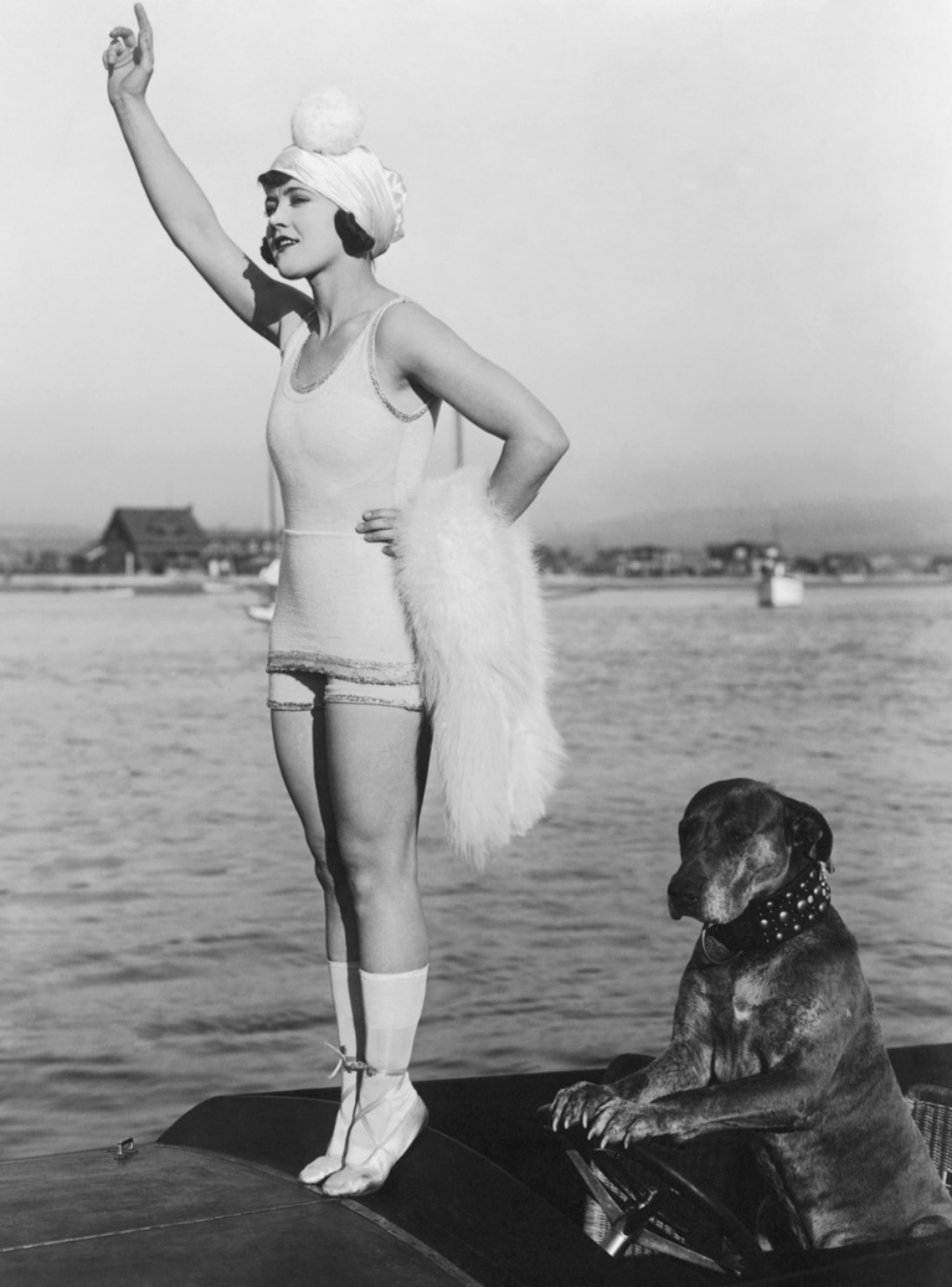 A woman in a vintage swimsuit, ballet shoes, and a cap with a pom-pom stands on a dock, raising her arm. A large dog wearing a spiked collar sits beside her, with water and buildings visible in the background.