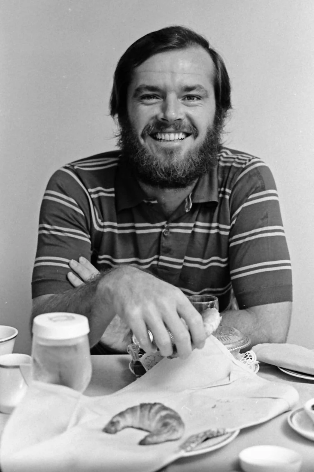 A man with a beard and striped shirt sits at a table, smiling at the camera. He holds a glass, with various breakfast items such as a croissant and a jar in front of him. The background is plain.