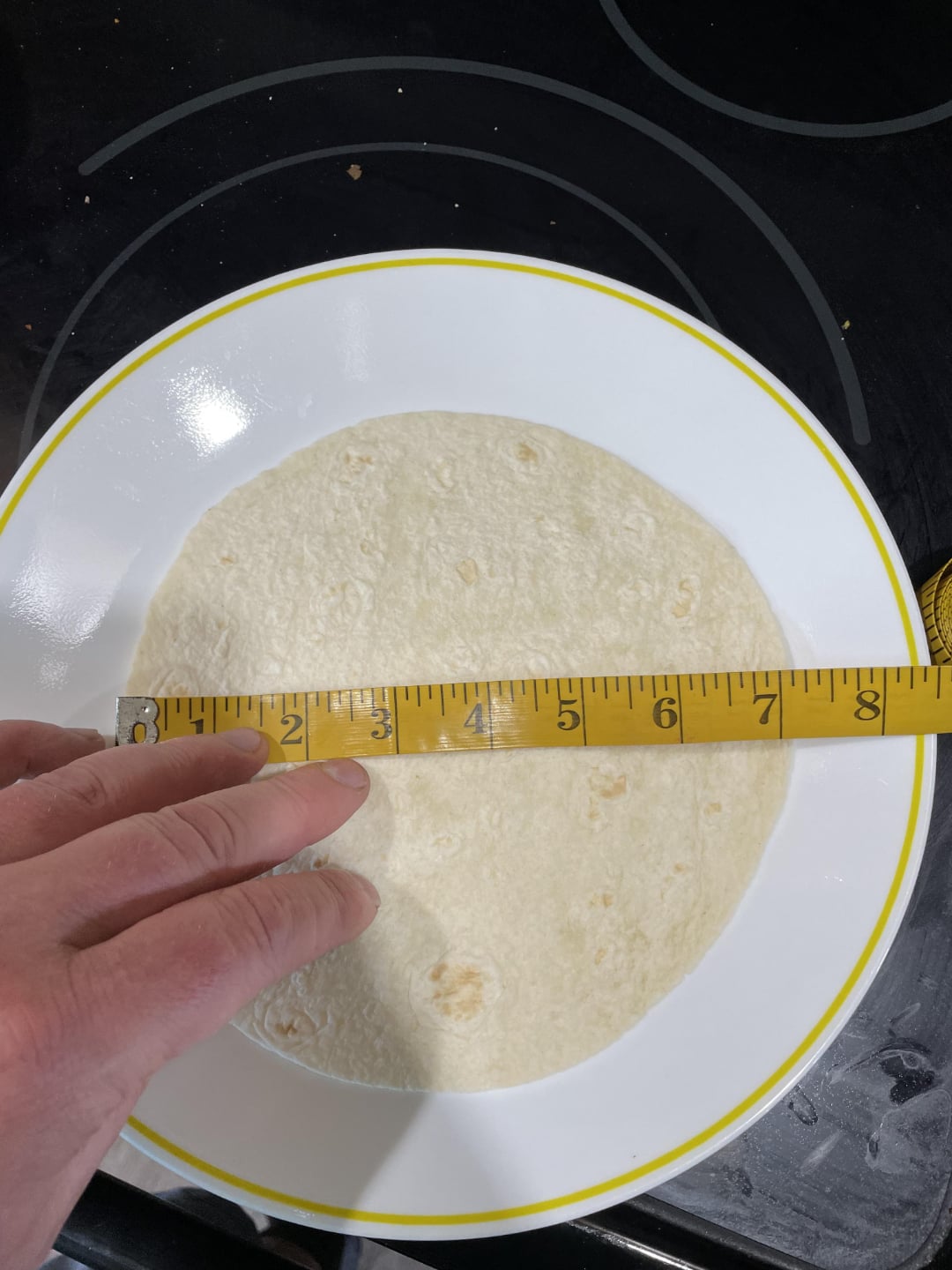 A hand holds a measuring tape across a flour tortilla placed on a white plate with a yellow rim, measuring the tortilla’s diameter at about 8 inches. The plate sits on a black stovetop.