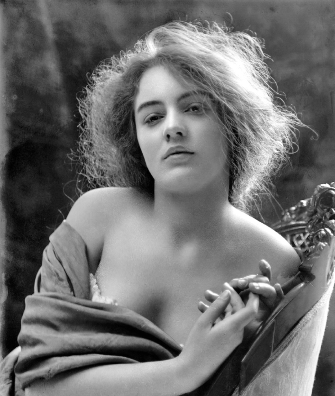 A black-and-white portrait of a woman with wavy, tousled hair, partially draped in fabric, gazing softly at the camera. Her hands are elegantly clasped, and she sits on an ornate chair.