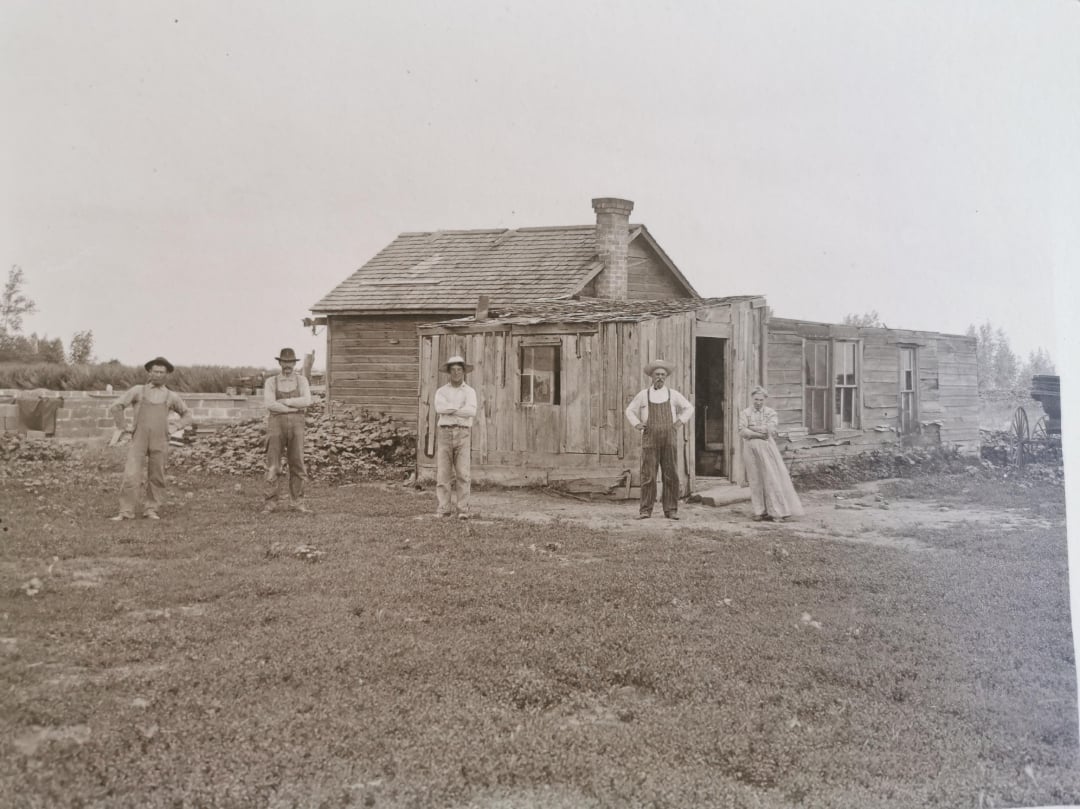 Five people stand in front of a small, weathered wooden house with a chimney, surrounded by grass and a few trees in the background; the scene is old-fashioned and in black and white.