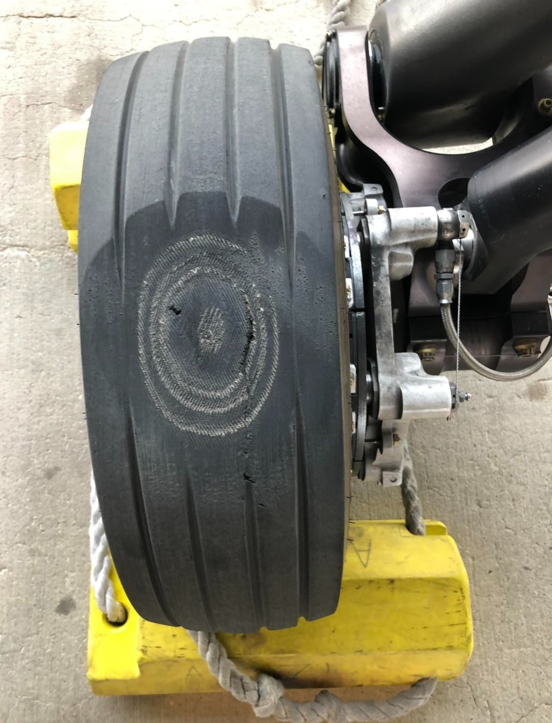 A close-up view of an aircraft tire with a severe flat spot, exposing underlying layers, positioned on a yellow wheel chock with part of the landing gear visible.