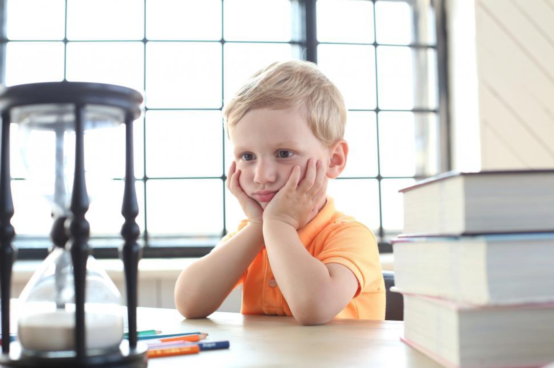 A young boy in an orange shirt sits at a table, resting his face in his hands, looking bored. An hourglass, colored pencils, and a stack of books are on the table in front of him. Large windows are in the background.