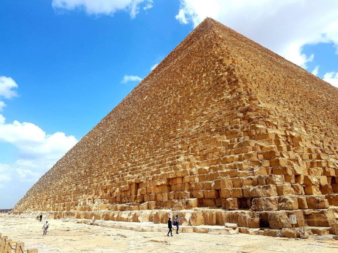 A large, ancient stone pyramid rises under a bright blue sky with scattered clouds. Several small figures of people stand near its massive base, highlighting the pyramid’s immense scale.