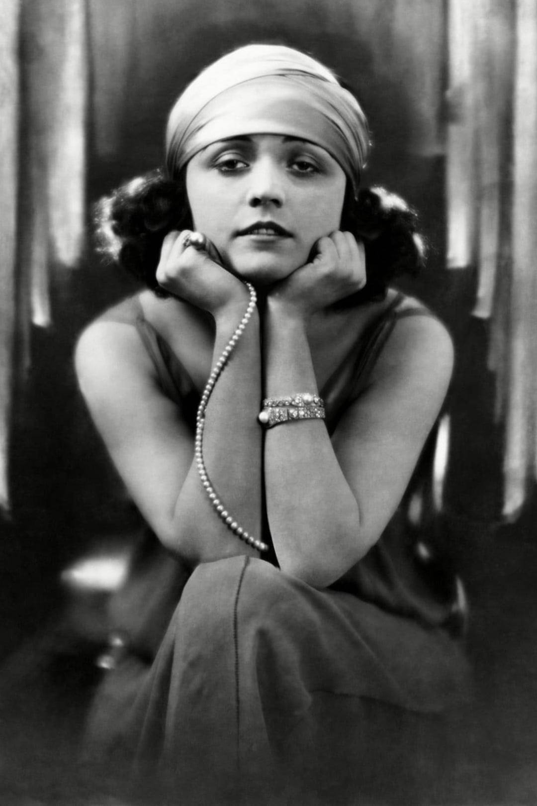 Black and white portrait of a woman in 1920s attire, wearing a headband, sleeveless dress, and pearl necklace, resting her chin on her hands and looking directly at the camera with a calm expression.
