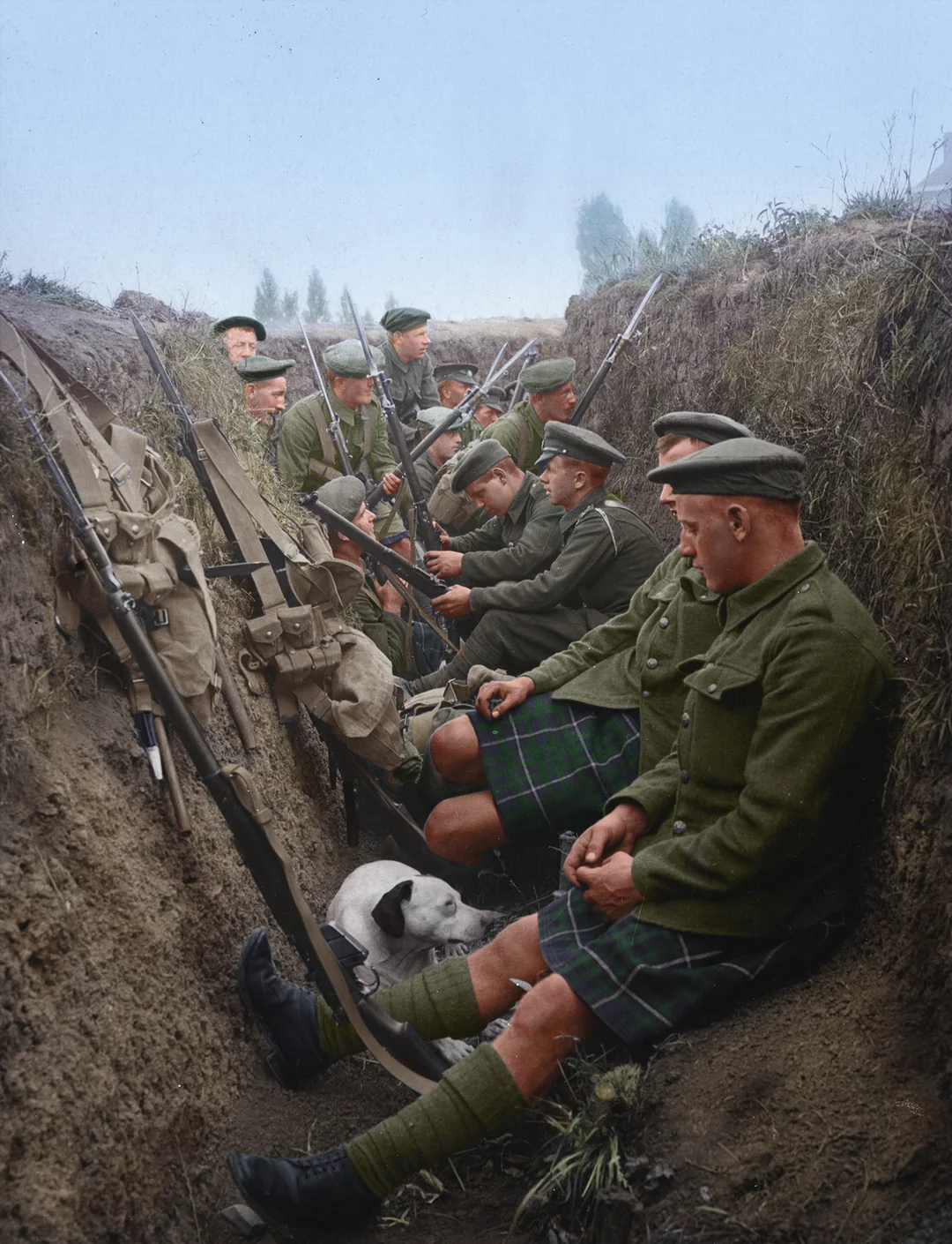 A group of World War I soldiers in uniform, some wearing kilts, sit in a narrow trench with rifles. A small dog sits among them. The mood appears calm as they wait and rest.