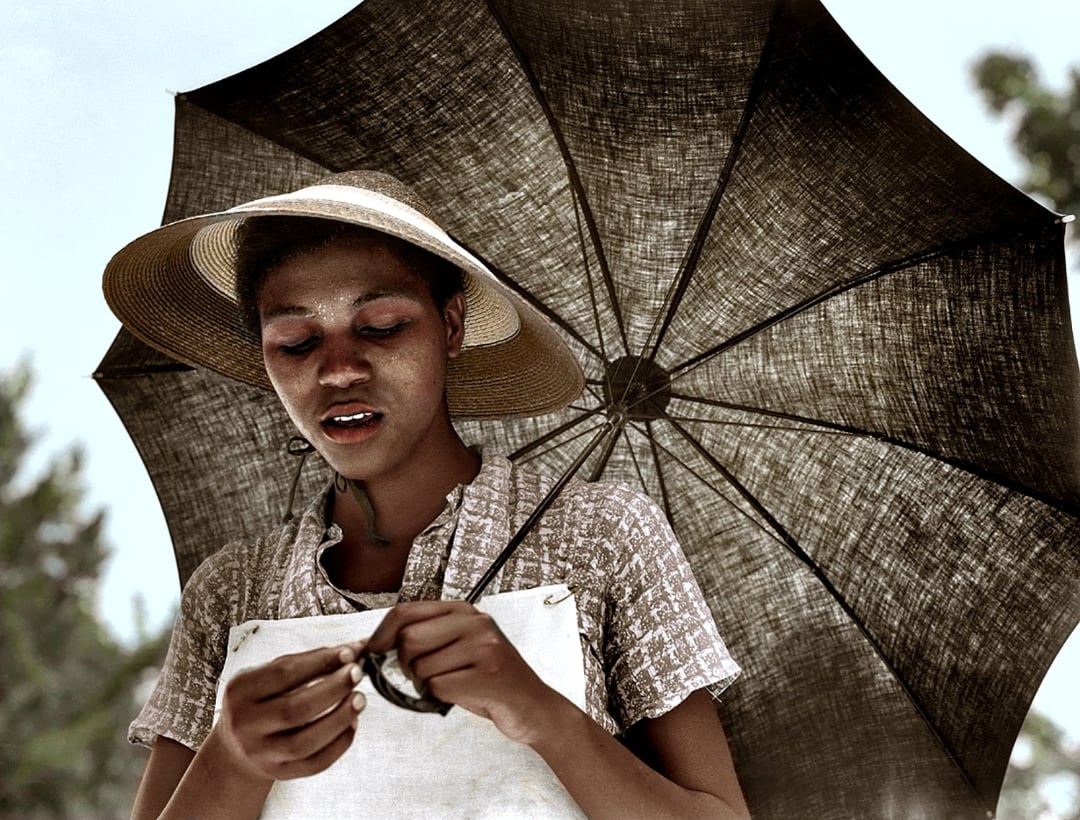 A woman wearing a straw hat and patterned dress stands under a large umbrella, looking down and working with her hands. The background is outdoors with trees visible.