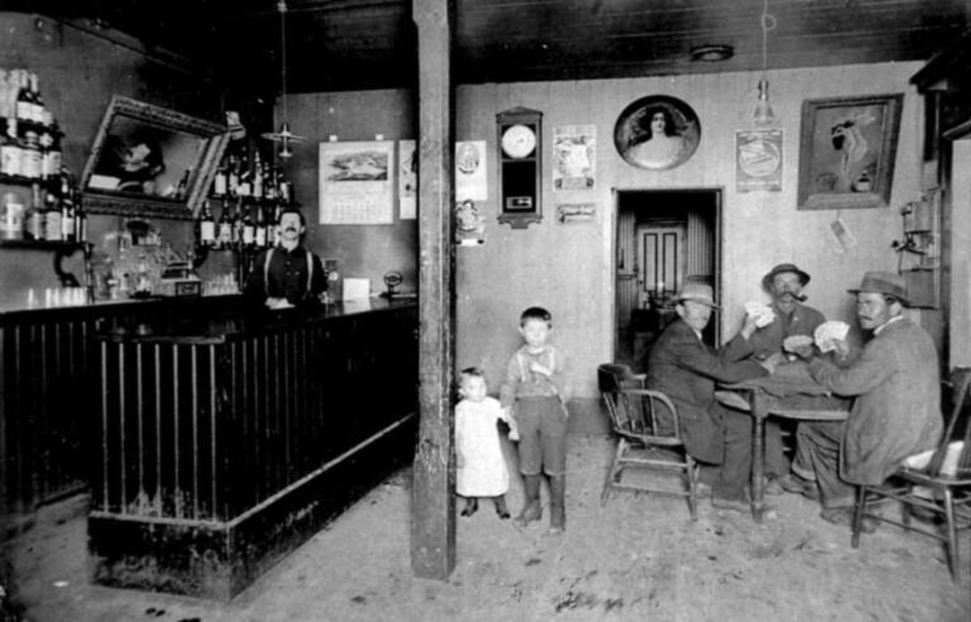 Black-and-white photo of an old-fashioned bar; a bartender stands behind the counter, two children stand near a wooden pillar, and four men sit at a table, one smoking, with bottles and pictures on the walls.