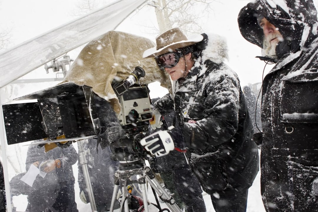 Two people in heavy winter gear operate a film camera outdoors in falling snow. One wears ski goggles and a cowboy hat, adjusting the camera, while the other stands nearby in a hooded coat. Snow covers their clothing and equipment.