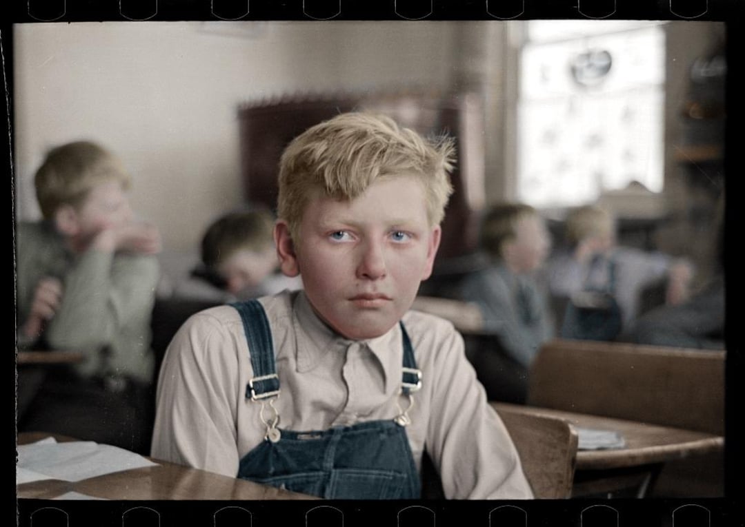 A blond boy in overalls sits at a desk in a classroom, looking serious. Other children and desks are blurred in the background, with light coming through a window. The image has a vintage, early 20th-century feel.