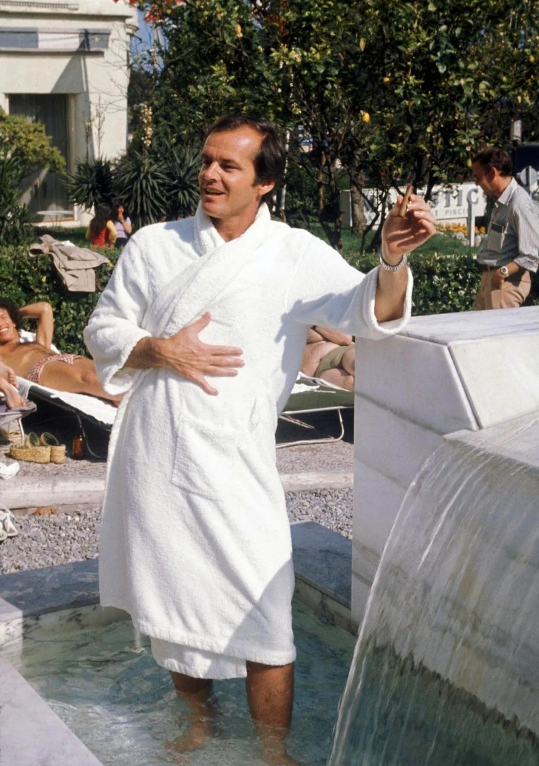 A man in a white bathrobe stands in a small fountain, smiling and gesturing with one hand. He is outdoors near a pool, with people relaxing on lounge chairs in the background on a sunny day.