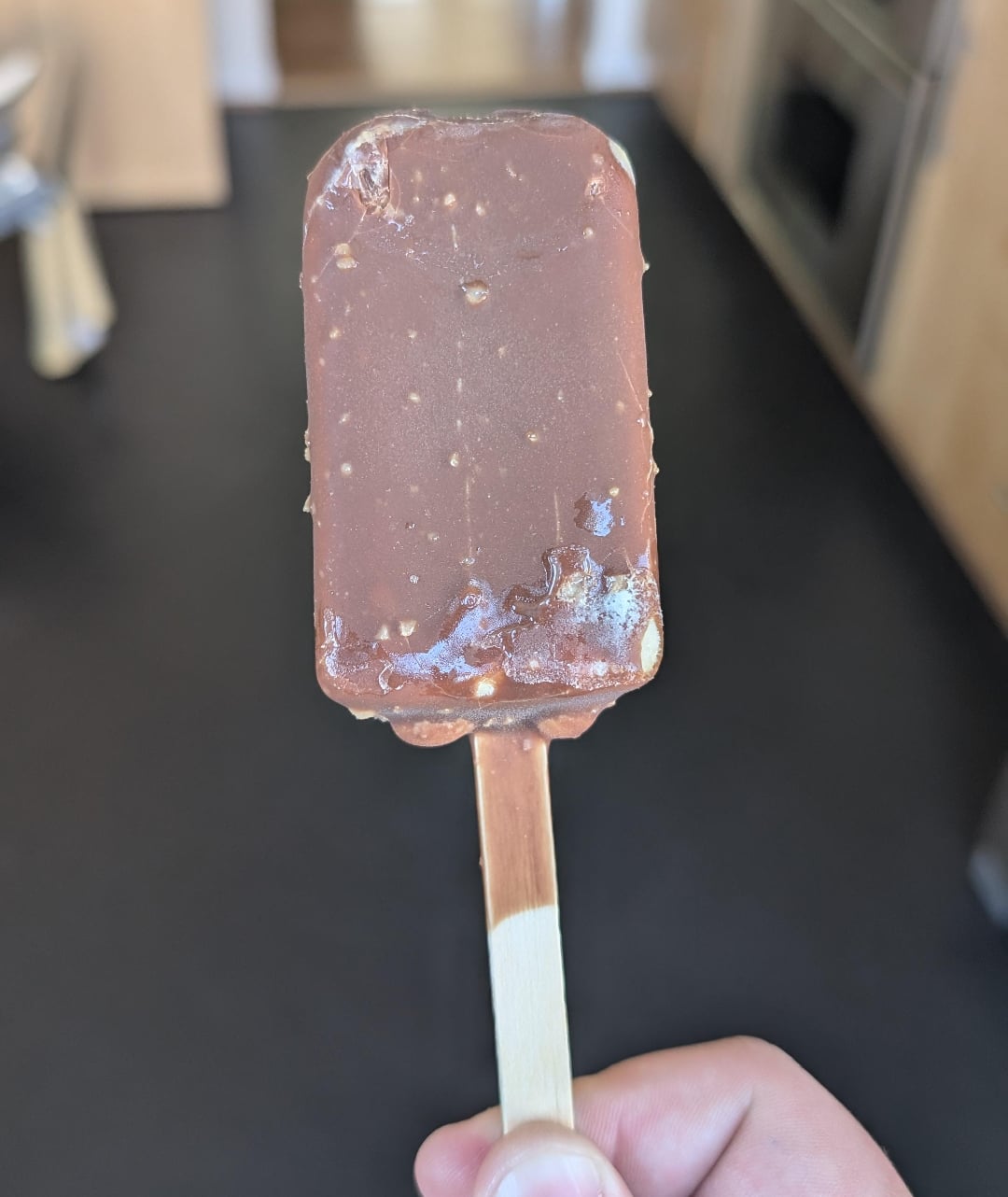 A hand holding a chocolate-covered ice cream bar on a wooden stick, with a kitchen background out of focus.
