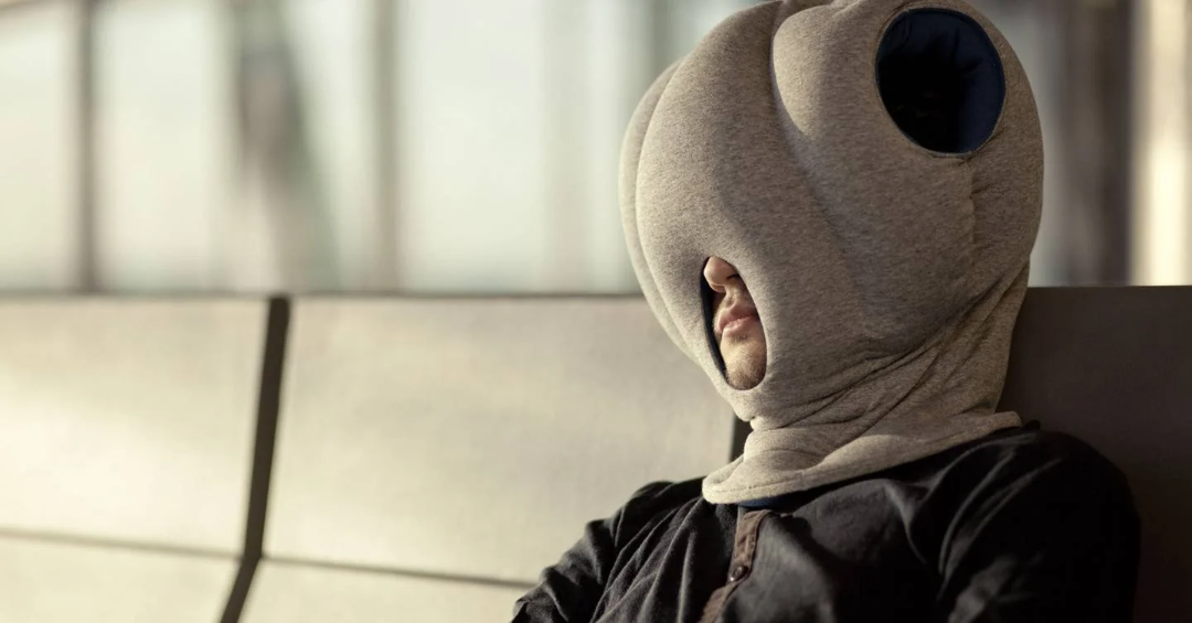 A person sits on an airport bench, wearing a large, padded, grey sleep hood that covers their entire head except for their nose and mouth, blocking out light and sound.