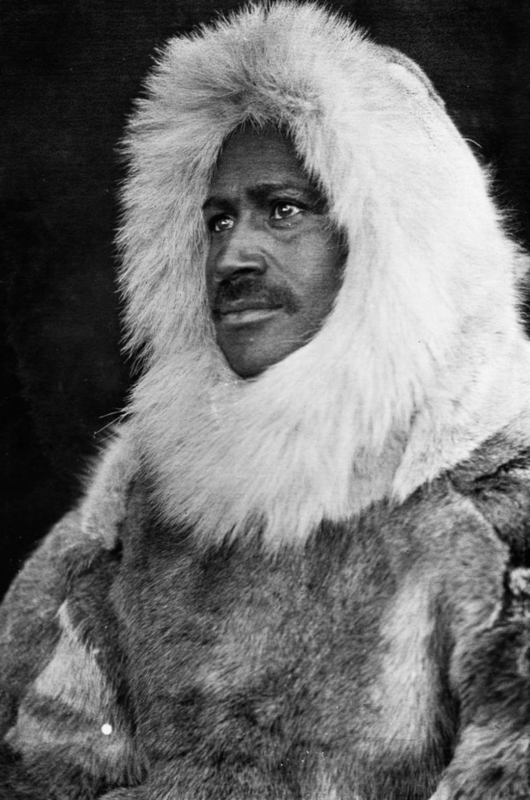 A person wearing a thick fur parka with a large hood, looking slightly to the side. The black-and-white photo gives a historical feel, and the fur clothing suggests a cold or Arctic environment.