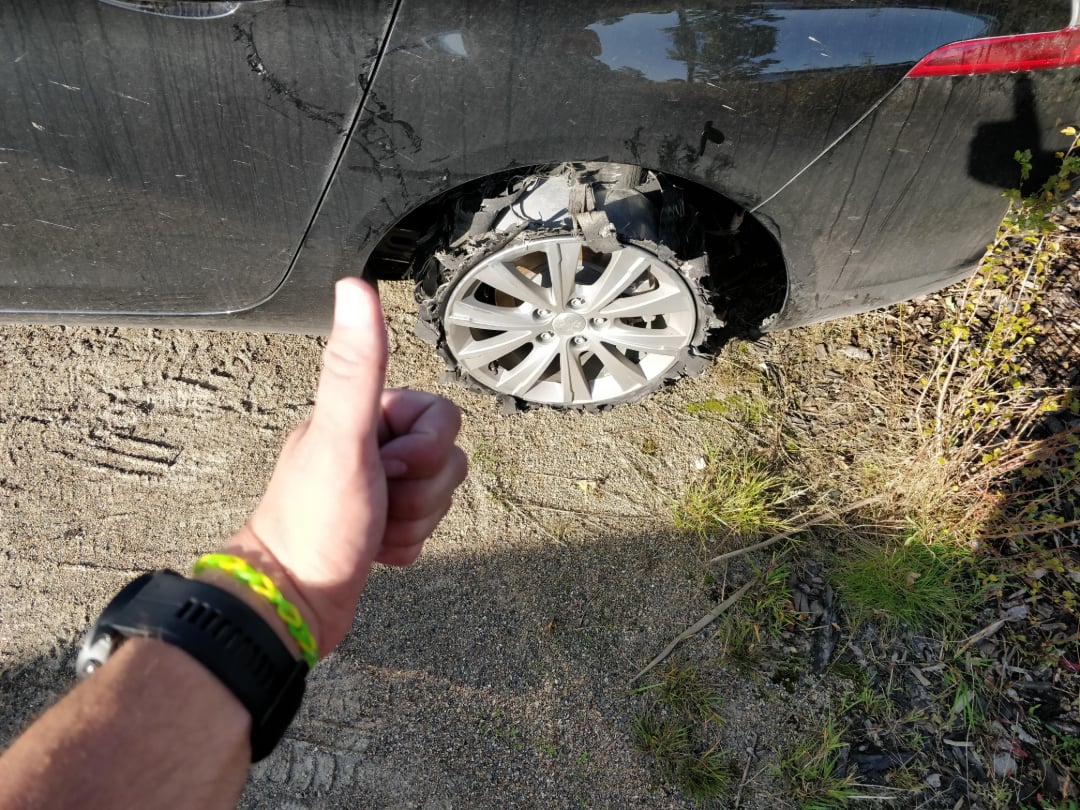 A person gives a thumbs-up in front of a car with a severely damaged rear tire, parked on a dirt surface. The tire is shredded, exposing the rim. The person's wristwatch and bracelets are visible.