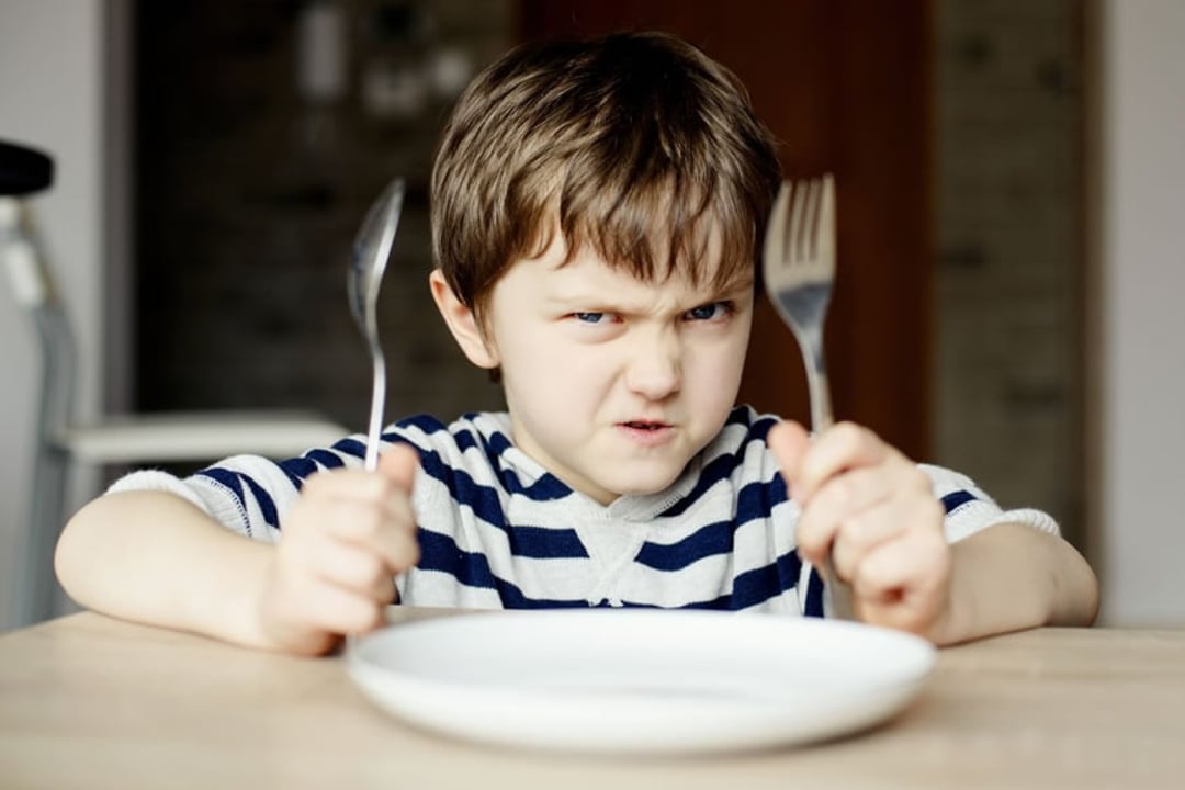 A young boy in a striped shirt sits at a table holding a fork and knife, looking impatient and frowning, with an empty white plate in front of him.