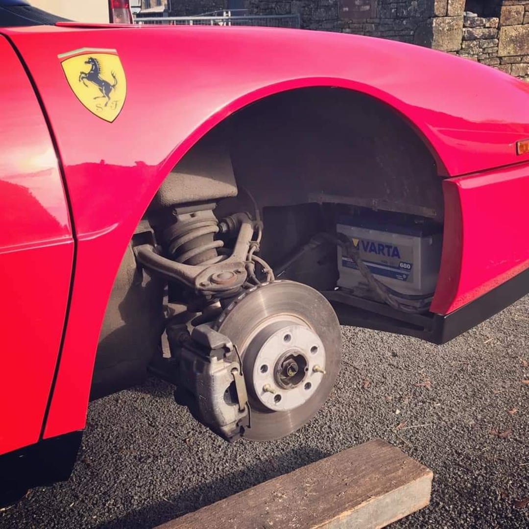 A red Ferrari with its front left wheel removed, exposing the brake disc, suspension, and a Varta battery inside the wheel well. The car is parked on a paved surface next to a stone wall.