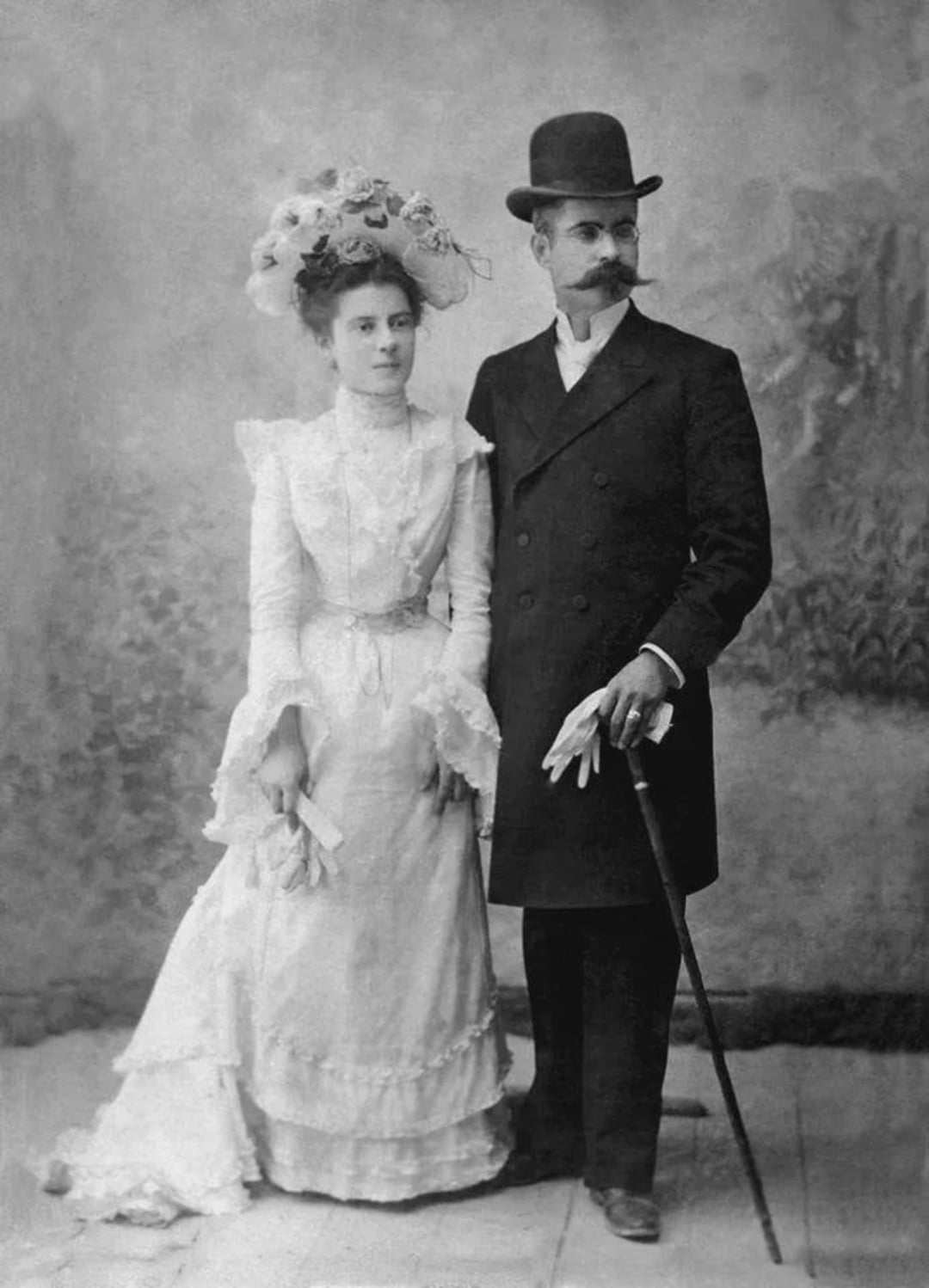 A vintage black-and-white photo of a woman in a long white dress and floral hat standing beside a man in a dark suit, bowler hat, and gloves, holding a cane. Both have neutral expressions and face the camera.