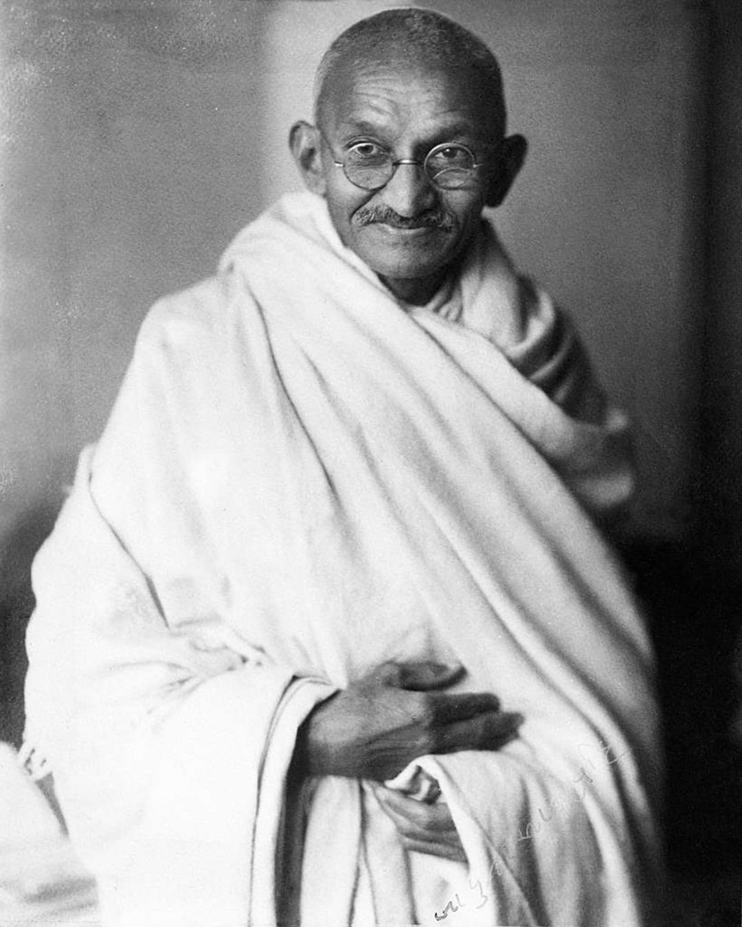 An elderly man with round glasses and a mustache, wrapped in a simple white cloth, stands facing the camera with a gentle smile and clasped hands. The background is plain and out of focus.