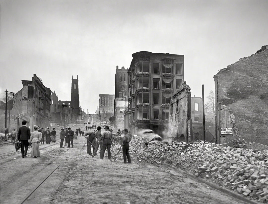 People walk along a rubble-strewn street flanked by damaged and partially destroyed buildings. Groups pause to observe the destruction. The scene appears somber and historic, with a tall church visible in the background.