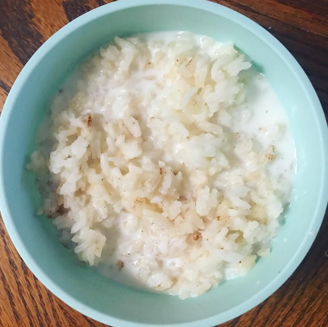 A light blue bowl containing rice mixed with milk, sprinkled with a small amount of cinnamon, sits on a wooden table.