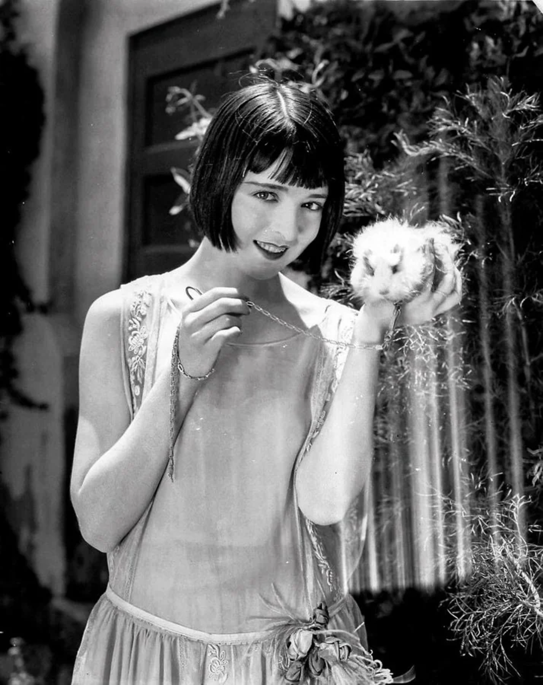 A woman with short dark hair and a sleeveless dress smiles while holding a fluffy white rabbit in one hand, standing outside near greenery and a door.