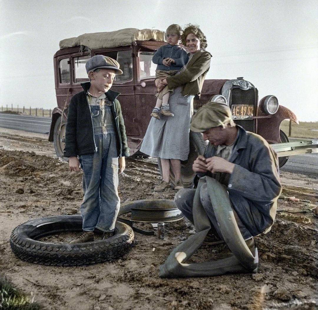 A family stands beside an old car on a dirt roadside; a man repairs a tire, a woman holds a child, and another child stands nearby. The scene appears to be from the early 20th century.