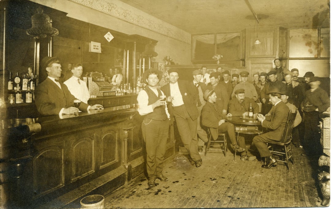A sepia-toned photo shows men in early 1900s attire gathered in a crowded bar, some standing at the counter and others seated around small tables, with bottles and glasses visible on the bar.