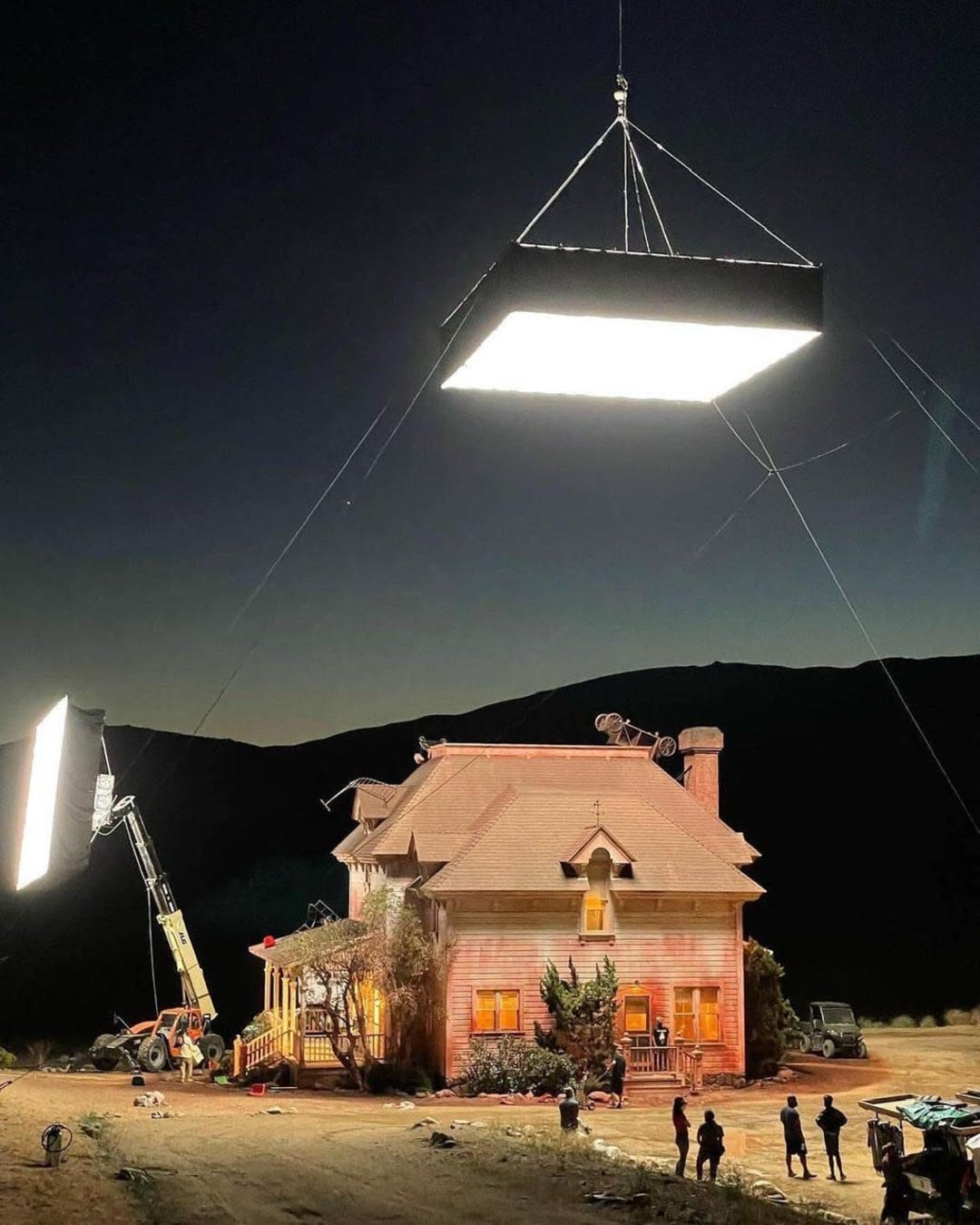 A large film light is suspended above a house on a movie set at night. Additional lighting equipment and crew members are visible around the house, which is illuminated against a dark, mountainous background.