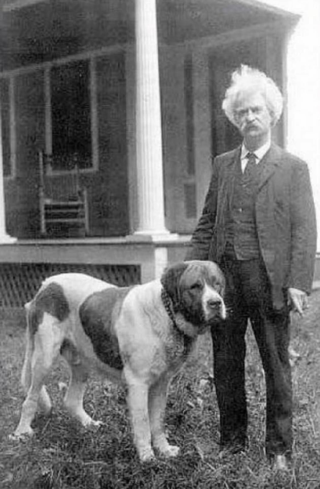 An older man with white hair and a mustache stands outside next to a large St. Bernard dog. They are in front of a porch with columns and a rocking chair visible in the background.