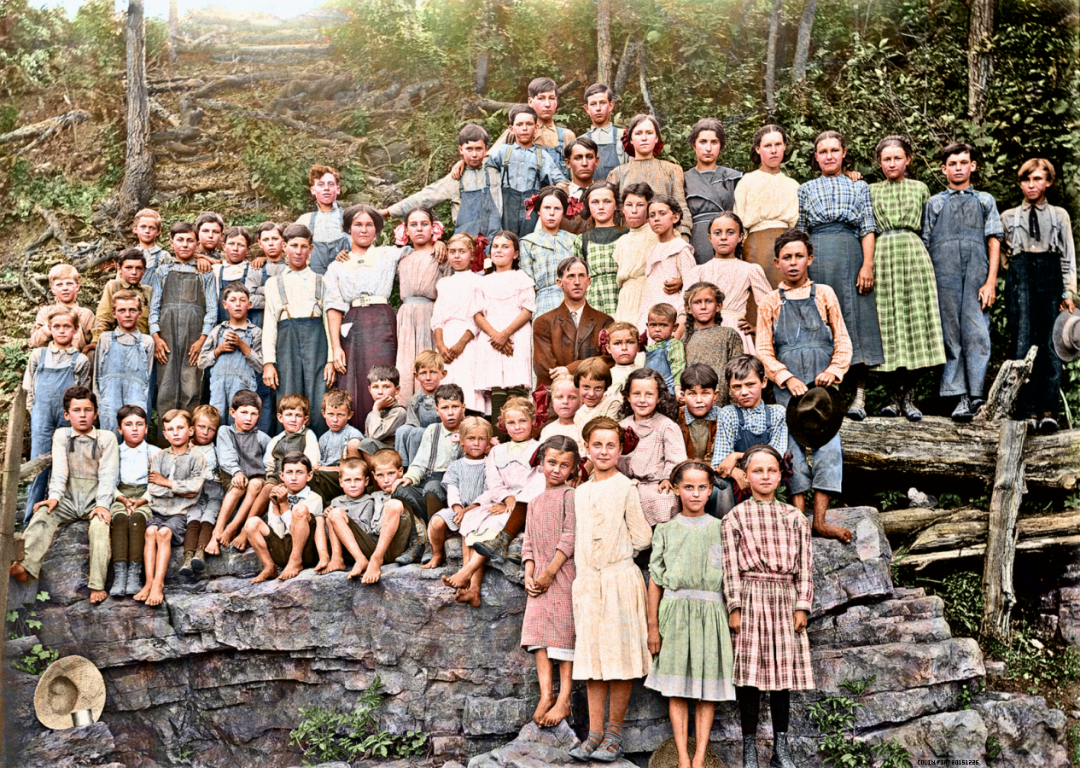A large group of children and a few adults pose outdoors on rocks and logs in front of a wooded area. The children wear old-fashioned clothing, including dresses and overalls, and appear to be from an earlier era.