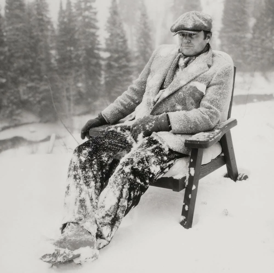 A man wearing a suit, gloves, and a flat cap sits in a wooden chair outdoors, covered in snow. Snow blankets his body and the ground around him, with snow-covered trees in the background.