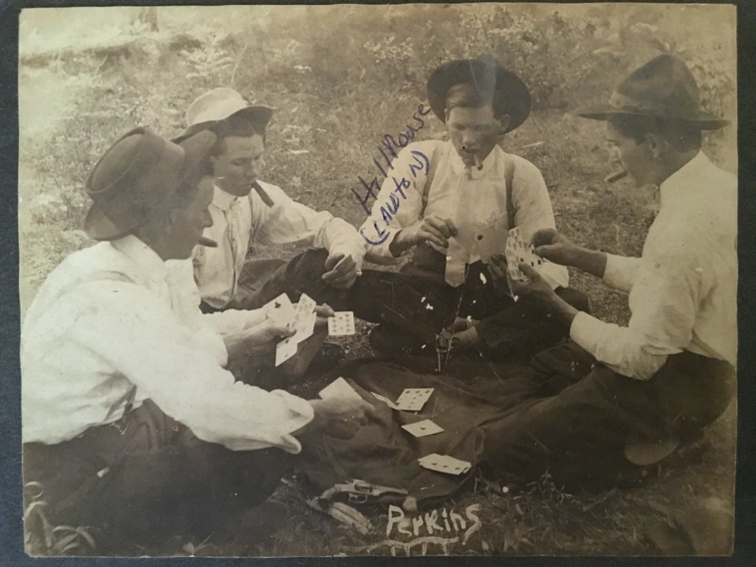 Four men in hats and old-fashioned clothing sit on a blanket outdoors, playing cards. Some cards are scattered on the ground. Two handwritten names, "Perkins" and "H. Williams (Lawton)," appear on the photo.