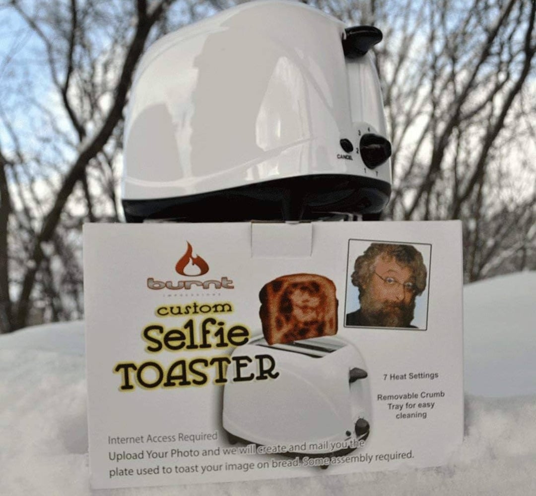 A white toaster sits on snow outdoors behind a product box that reads "burnt custom Selfie TOASTER" and shows a piece of toast with a bearded man's face toasted on it, along with an image of the man. Bare trees are in the background.