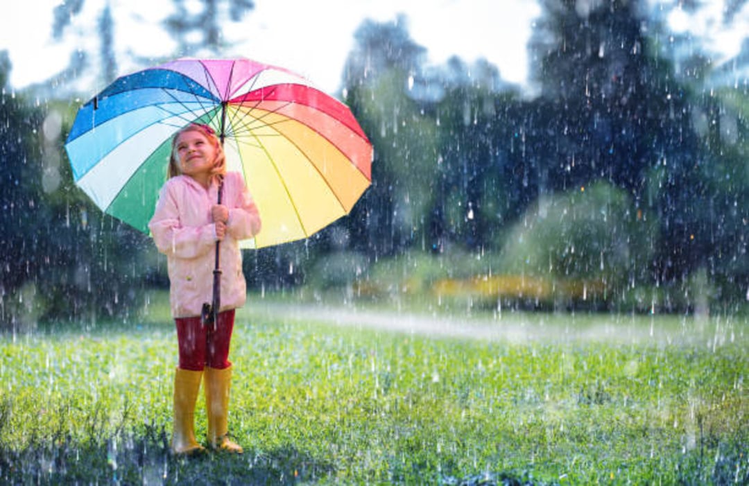 A young child stands on grass in the rain holding a large rainbow-colored umbrella, smiling and looking up. The child is wearing a light-colored jacket, red pants, and yellow rain boots.