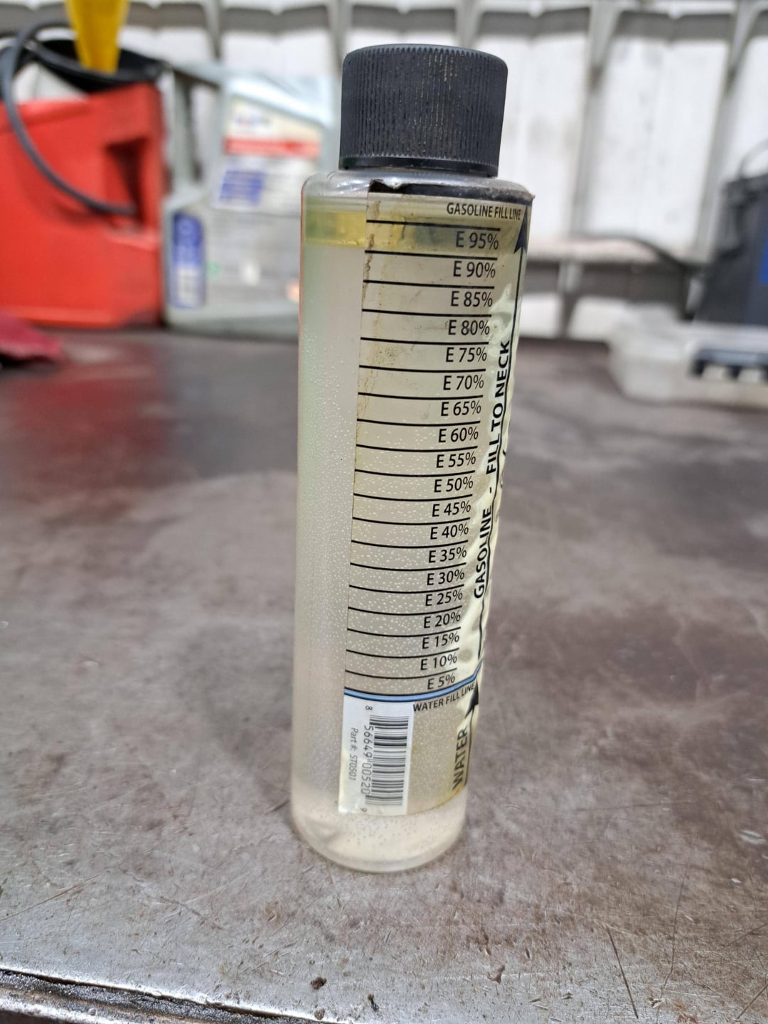 A clear plastic bottle with a black cap is partially filled with liquid, showing measurement markings labeled E 95% to E 10% and "GASOLINE FAILURE" on the side, placed on a metal surface in a workshop setting.