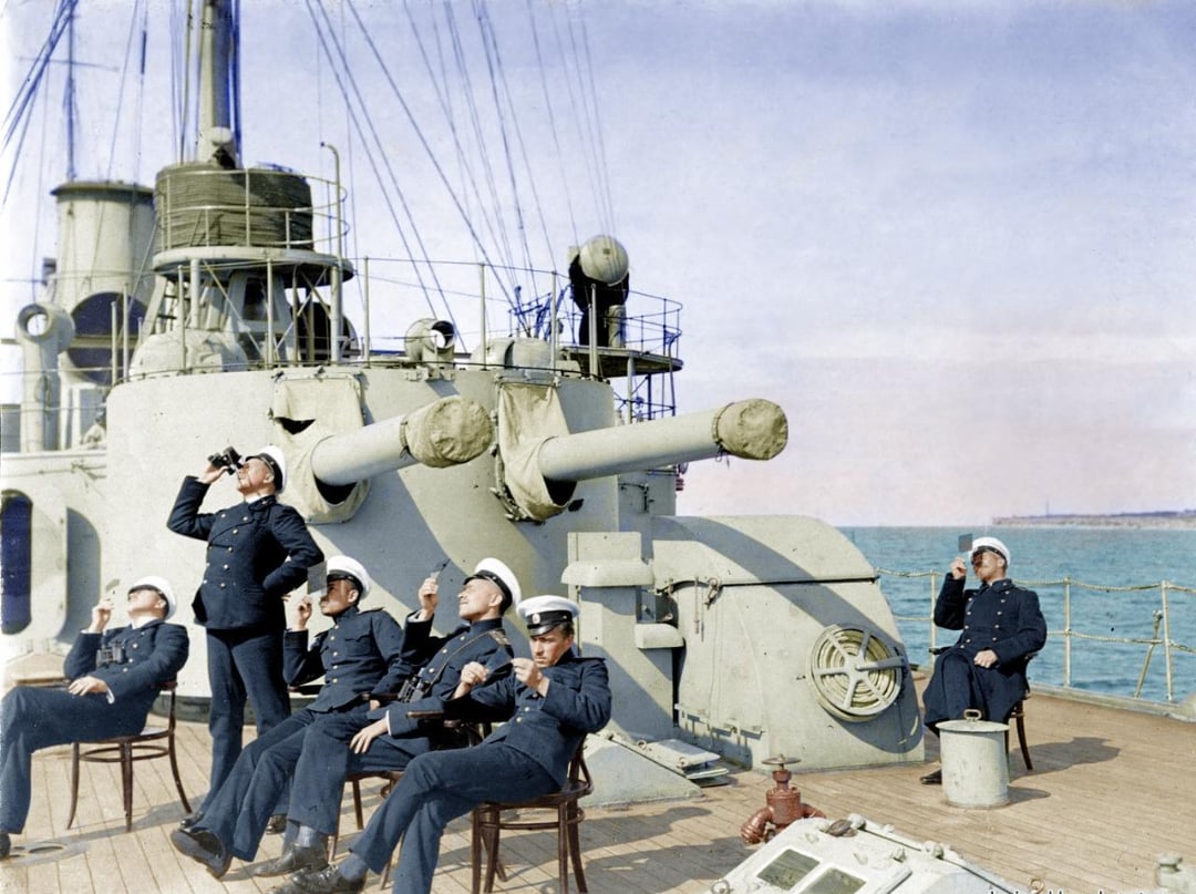 Six naval officers in dark uniforms relax and drink on the deck of a ship near large cannons, with blue sea and sky visible in the background. Some are seated on chairs while others stand or sit on different parts of the deck.