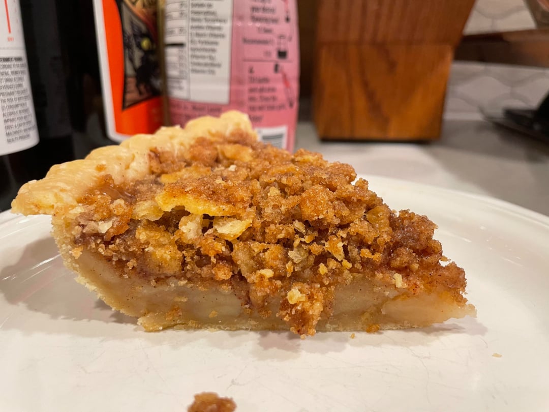A close-up of a slice of pie with a crumbly, golden-brown topping and a light, custard-like filling, served on a white plate with various bottles and a wooden block in the background.