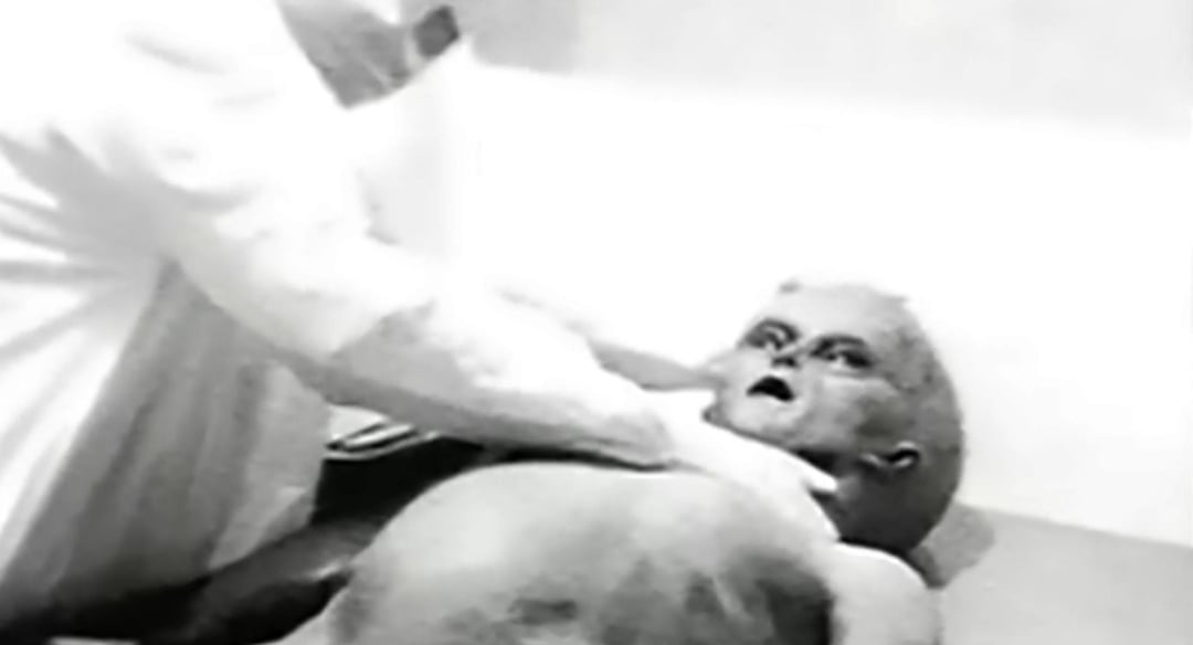 A person in a protective suit examines a figure resembling an alien lying on a table in a black and white setting. The figure has a large head, dark eyes, and an open mouth.