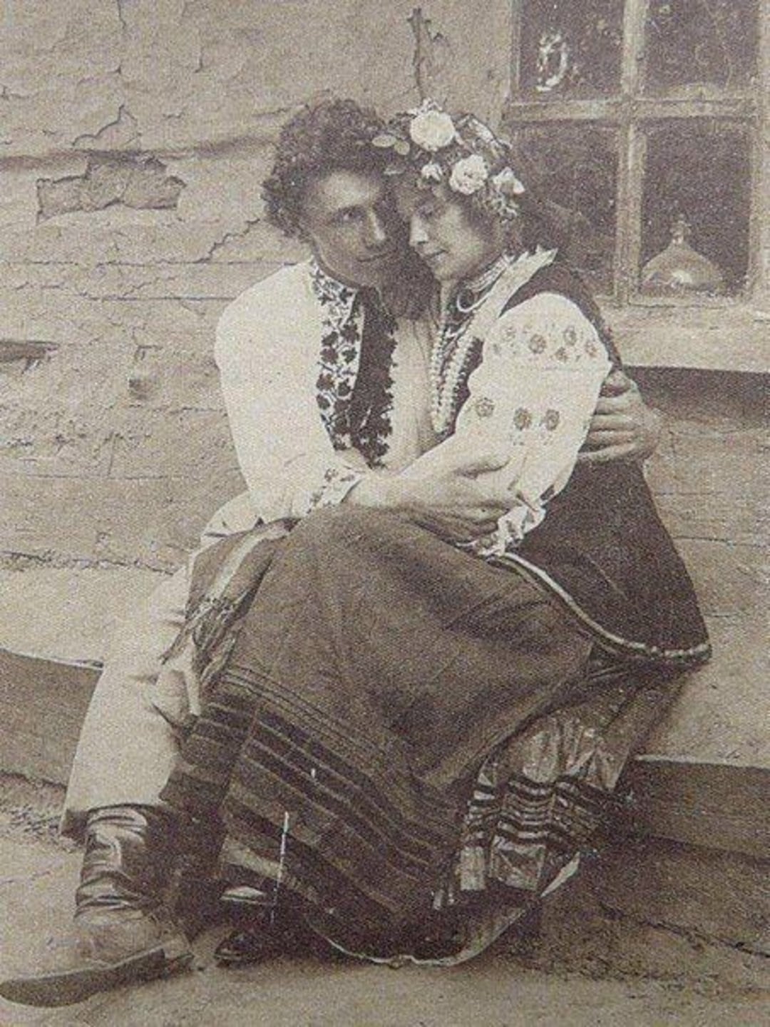 A vintage photo shows a young couple in traditional folk clothing sitting closely together; the man embraces the woman, who wears a floral headpiece, outside a rustic building with a wooden window.