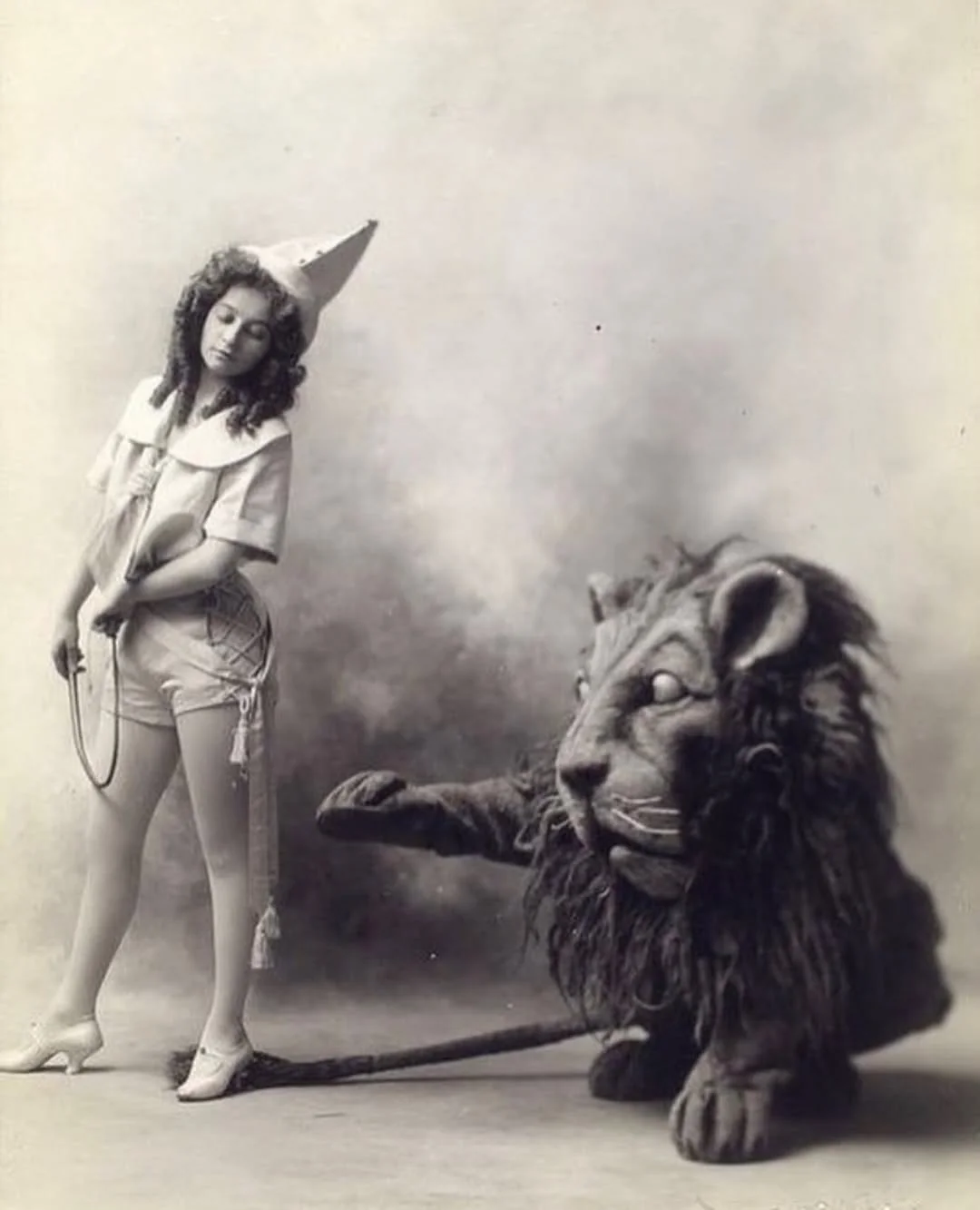 A young girl in a vintage costume with a pointed hat stands holding a whip next to a large, lifelike lion costume or puppet. The setting is a simple studio with a plain backdrop.